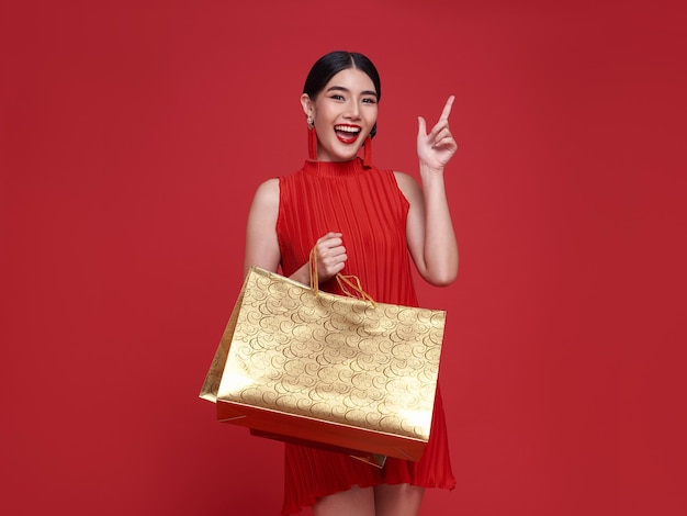 Sorrindo linda mulher asiática segurando sacolas de compras e apontando para copiar o espaço isolado em fundo vermelho para o conceito de venda de ano novo chinês.
