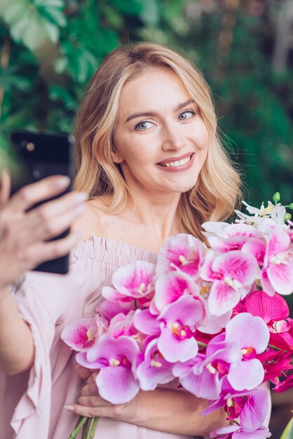 Sorrindo, loiro, mulher jovem, segurando, orquídea, mão, levando, retrato próprio, de, telefone móvel