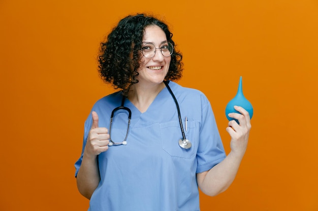 Foto grátis sorrindo médica de meia idade usando óculos uniformes e estetoscópio em volta do pescoço mostrando enema olhando para a câmera mostrando o polegar isolado em fundo laranja