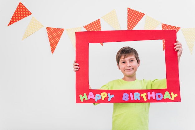 Sorrindo, menino, segurando, quadro, com, feliz aniversário, armação, contra, parede, decorado, com, bunting