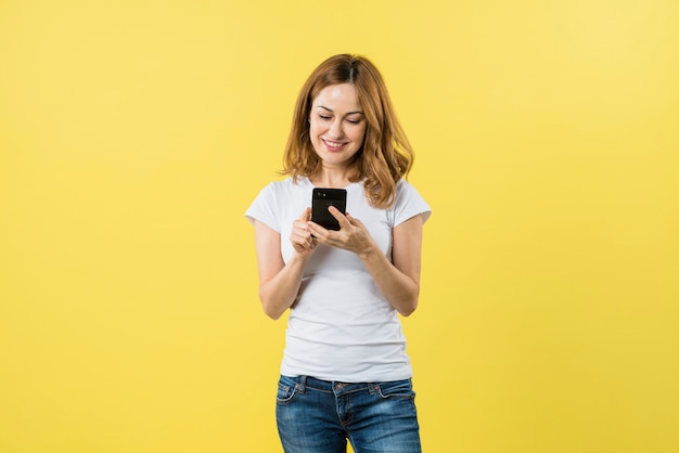 Sorrindo mensagens de mensagens de texto jovem loira no telefone móvel contra um fundo amarelo