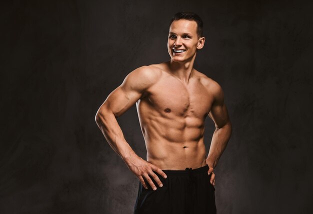 Sorrindo modelo de fitness jovem bonito com corpo musculoso posando no estúdio em um fundo escuro.