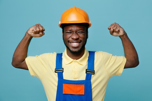 Sorrindo mostrando forte gesto jovem construtor americano africano de uniforme isolado em fundo azul