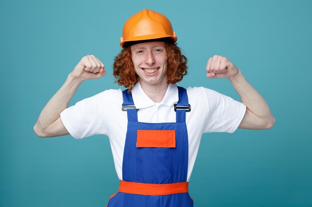 Sorrindo mostrando forte gesto jovem construtor de uniforme isolado em fundo azul