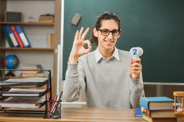 sorrindo mostrando o gesto ok professor de óculos sentado na mesa segurando números de matemática com ferramentas escolares em sala de aula
