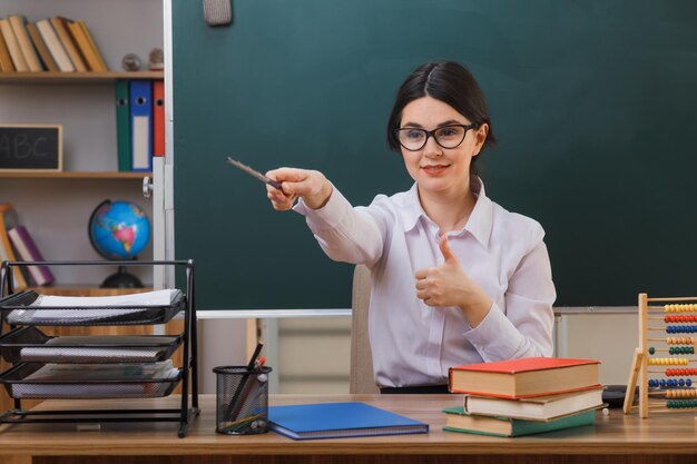 sorrindo mostrando os polegares para cima jovem professora usando óculos aponta ao lado com ponteiro sentado na mesa com ferramentas escolares em sala de aula