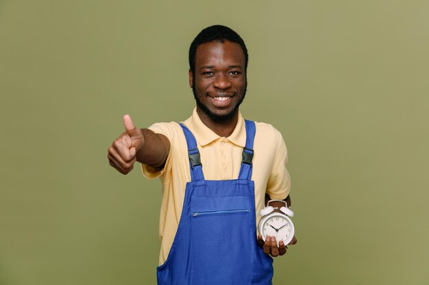 Sorrindo, mostrando os polegares para cima, segurando o despertador, jovem limpador afro-americano de uniforme com luvas isoladas em fundo verde