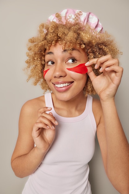 Foto grátis sorrindo muito jovem coloca manchas vermelhas de beleza sob os olhos passa por procedimentos de cuidados com a pele usa chapéu de banho e camiseta casual isolada sobre fundo cinza conceito de bem-estar e rejuvenescimento