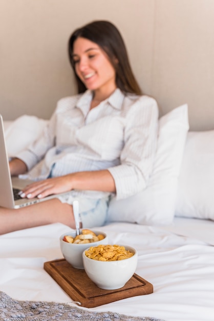 Sorrindo, mulher jovem, usando computador portátil, com, cornflakes, e, salada fruta, cama