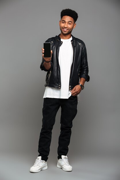 sorrindo na moda cara Africano em pé com a mão no bolso enquanto mostra a tela do celular em branco, olhando