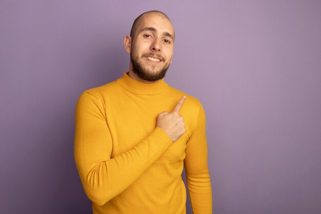 Sorrindo, olhando para a frente, jovem bonito apontando para o lado isolado em roxo com espaço de cópia