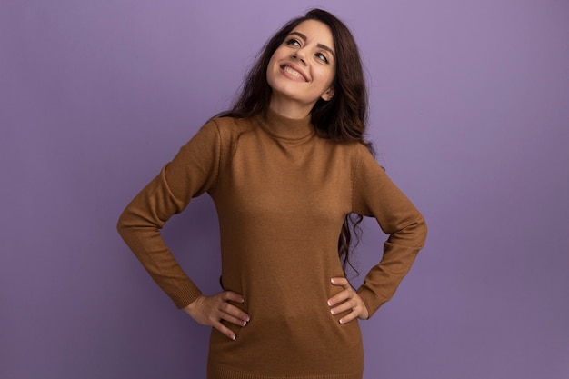 Foto grátis sorrindo, olhando para o lado da jovem linda, vestindo uma blusa de gola alta marrom, colocando as mãos no quadril isoladas na parede roxa