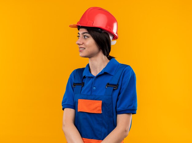 Sorrindo, olhando para o lado jovem construtora de uniforme isolado na parede amarela com espaço de cópia