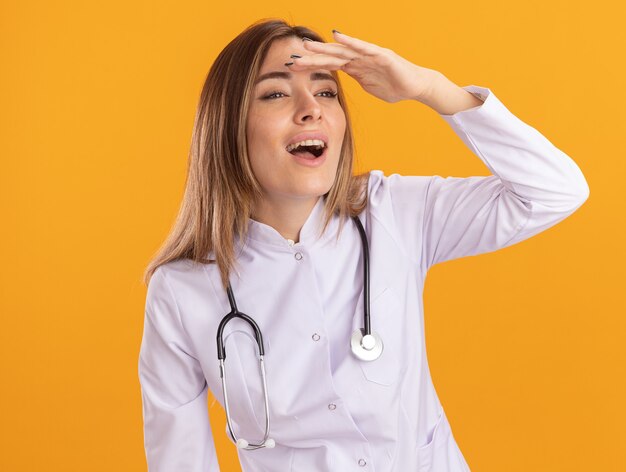 Sorrindo, olhando para o lado, jovem médica vestindo bata médica com estetoscópio, olhando para a distância com a mão isolada na parede amarela