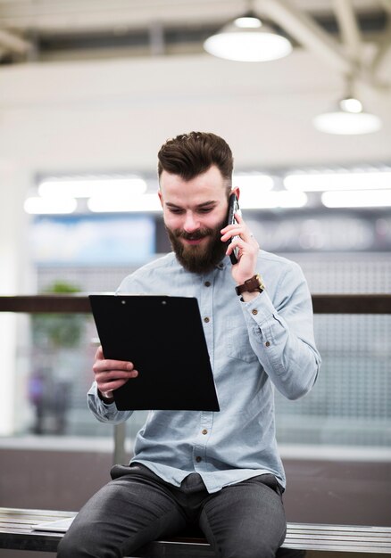 Sorrindo, retrato, de, um, jovem, homem negócios, olhar, prancheta, falando telefone móvel