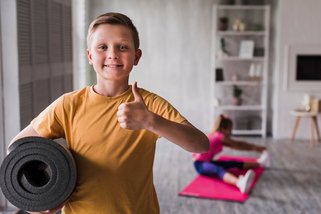 Foto grátis sorrindo, retrato, de, um, menino, segurando, rolado, esteira exercício, mostrando, polegares cima, sinal