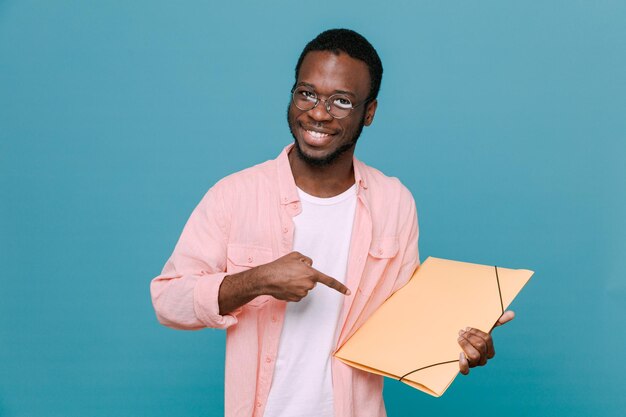 Sorrindo segurando e aponta para pasta jovem afro-americano isolado em fundo azul