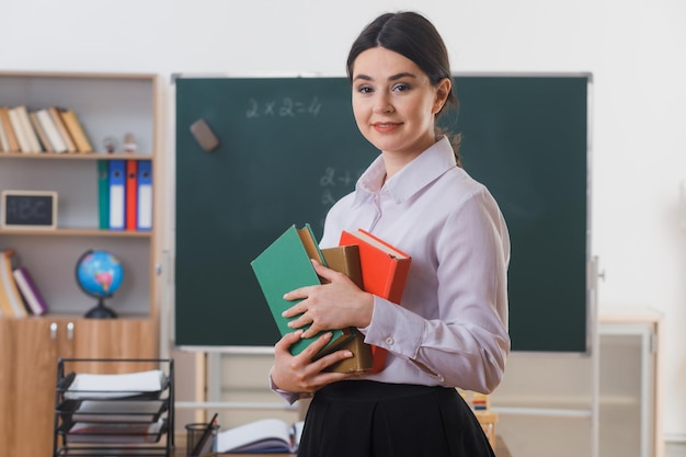 Foto grátis sorrindo segurando livros jovem professor em frente ao quadro-negro em sala de aula