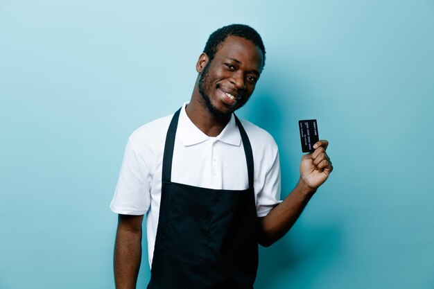 Sorrindo segurando o cartão jovem barbeiro americano africano de uniforme isolado em fundo azul