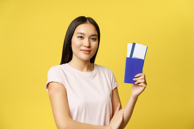 Sorrindo vacinado de uma menina asiática cobiçosa, mostrando passaporte com passagens, recebendo vacina contra coronavírus para viajar para o exterior, fundo amarelo