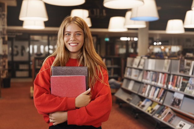 Sorriso alegre, garota de leitura manual segurando livros, em pé na biblioteca ou livraria.
