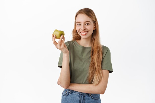 Sorriso branco. Mulher jovem feliz comendo maçã verde, mostrando os dentes perfeitos e parecendo satisfeita com a câmera, vestindo uma camiseta contra um fundo branco