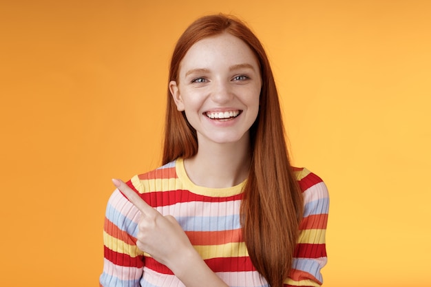 Sorriso carismático feliz, jovem ruiva, olhos azuis, sorriso sincero e carinhoso, rindo amplamente se divertindo, discutindo concerto hilário apontando o canto superior esquerdo mostrando um produto incrível, fundo laranja