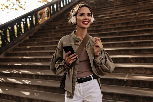 Sorrisos de mulher feliz com cabelo curto e lábios vermelhos em fones de ouvido. mulher de jaqueta e calças leves segura o telefone ao ar livre.