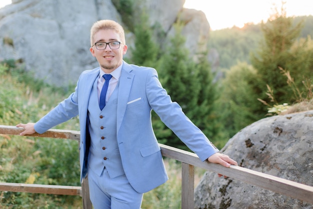 Sorriu jovem elegante vestido de terno azul elegante e óculos está de pé perto de enormes pedras