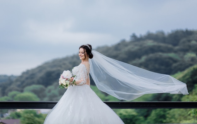 Sorriu muito caucasiana noiva com véu longo ao ar livre, segurando o buquê de casamento