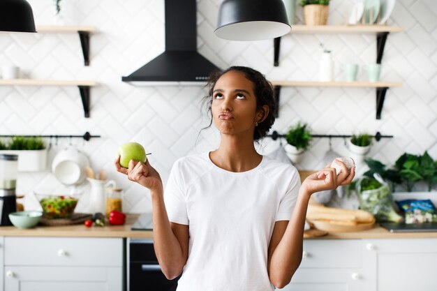 Sorriu mulata atraente está pensando em uma maçã com rosto hilariante e olhando para o topo na cozinha moderna branca