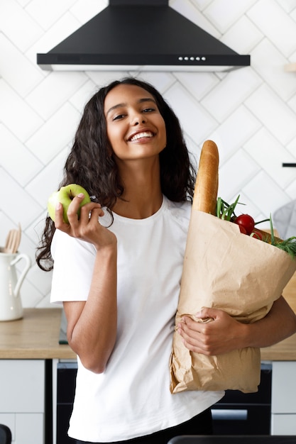 Sorriu mulata bonita está segurando o pacote cheio com comida em uma mão e maçã em outra na moderna cozinha branca