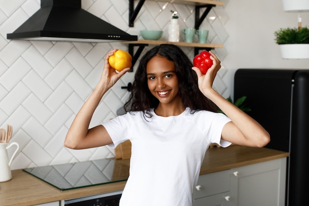 Sorriu mulata com cabelo solto está segurando pimentões vermelhos e amarelos nas mãos perto da cabeça na cozinha moderna, vestida com camiseta branca