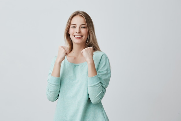 Sorte, jovem funcionária, regozijando-se com sucesso no trabalho, sorrindo amplamente, mantendo os punhos cerrados. Mulher loira bonita camisola azul clara, sentindo-se feliz e animado posando