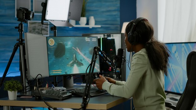 Streamer de mulher negra jogando videogames de tiro espacial com joystick, conversando com colegas de equipe no chat aberto de streaming. Cyber desempenho em computador poderoso RGB em sala de jogos usando equipamento profissional