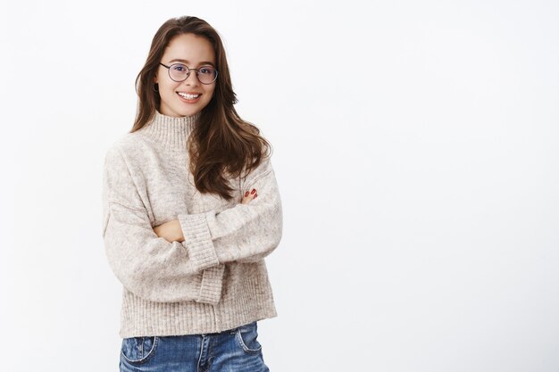 Studi foto de elegante criativo e feliz jovem europeu em copos com sorriso perfeito de mãos cruzadas sobre o peito em pose confiante e otimista, vestindo uma camisola quente num dia frio de inverno.