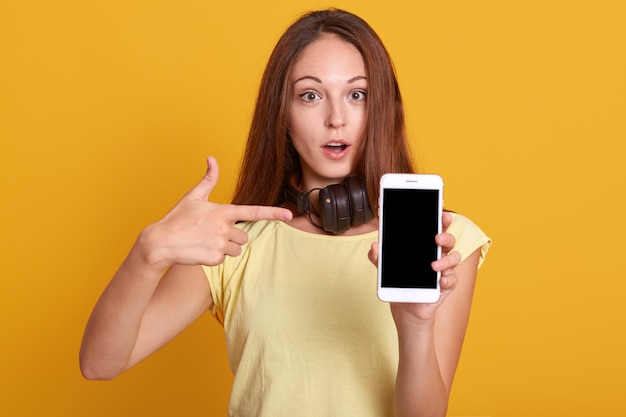 Studio sho de mulher adorável, mostrando o telefone com tela em branco e apontando com o dedo indicador