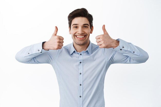 Super bom. Homem bonito sorridente mostrando os polegares para cima em aprovação, gosto e concorda, elogia o bom trabalho, parado satisfeito contra um fundo branco