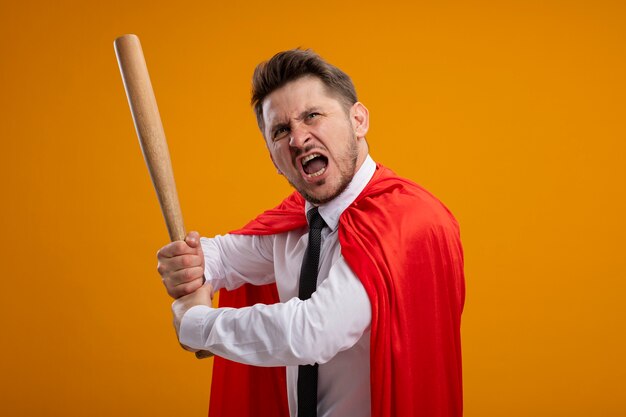 Super-herói empresário com capa vermelha balançando o taco de beisebol gritando com expressão agressiva em pé sobre a parede laranja