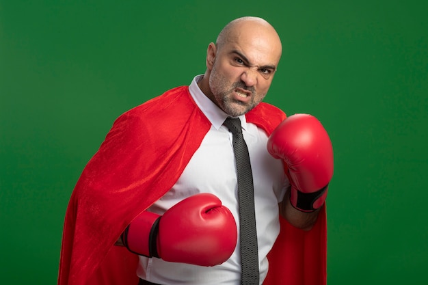 Super-herói empresário com capa vermelha e luvas de boxe olhando para frente com cara de raiva pronto para lutar, posando como um lutador em pé sobre a parede verde