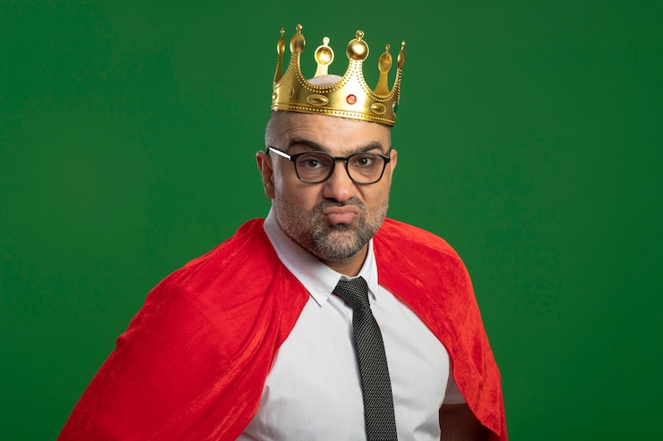 Foto grátis super-herói empresário com capa vermelha e óculos usando coroa, olhando para a frente, satisfeito consigo mesmo em pé sobre a parede verde