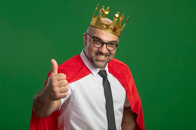 Foto grátis super-herói empresário com capa vermelha e óculos usando coroa, olhando para a frente, sorrindo com uma cara feliz mostrando os polegares para cima em pé sobre a parede verde