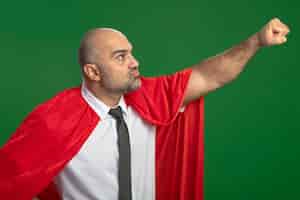 Foto grátis super-herói empresário com capa vermelha olhando para o lado, mantendo o braço em gesto voador pronto para ajudar, parecendo confiante em pé sobre a parede verde