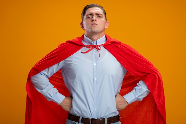 Super-herói empresário com capa vermelha, parecendo confiante, com uma expressão séria e os braços na cintura, prontos para ajudar em pé sobre um fundo laranja
