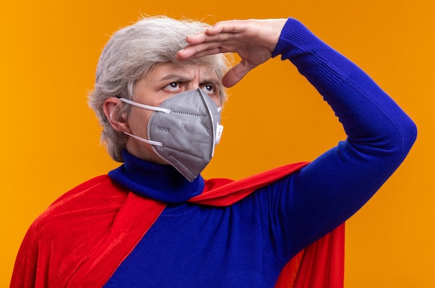 Foto grátis super-heroína sênior usando capa vermelha e máscara protetora facial, olhando para longe com uma mão sobre a cabeça em pé sobre um fundo laranja