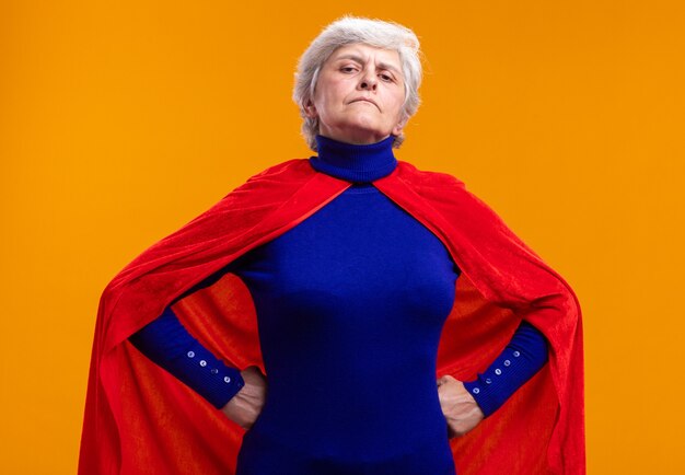 Foto grátis super-heroína sênior usando capa vermelha, olhando para a câmera com expressão confiante, com os braços na altura do quadril em pé sobre a laranja