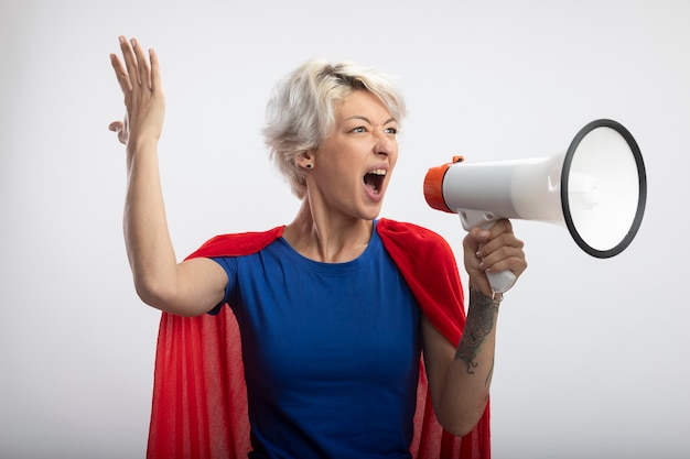 Supermulher irritada com capa vermelha fica de pé com a mão levantada e grita no alto-falante isolado na parede branca