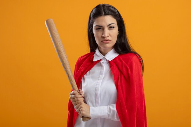 Supermulher jovem confiante segurando um taco de beisebol, olhando para a frente, isolado na parede laranja