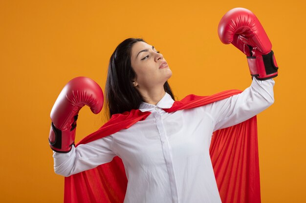 Supermulher jovem confiante usando luvas de caixa fazendo um gesto forte, olhando para a mão dela isolada na parede laranja