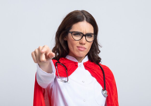 Supermulher séria em uniforme de médico com capa vermelha e estetoscópio em óculos ópticos aponta na frente isolado na parede branca